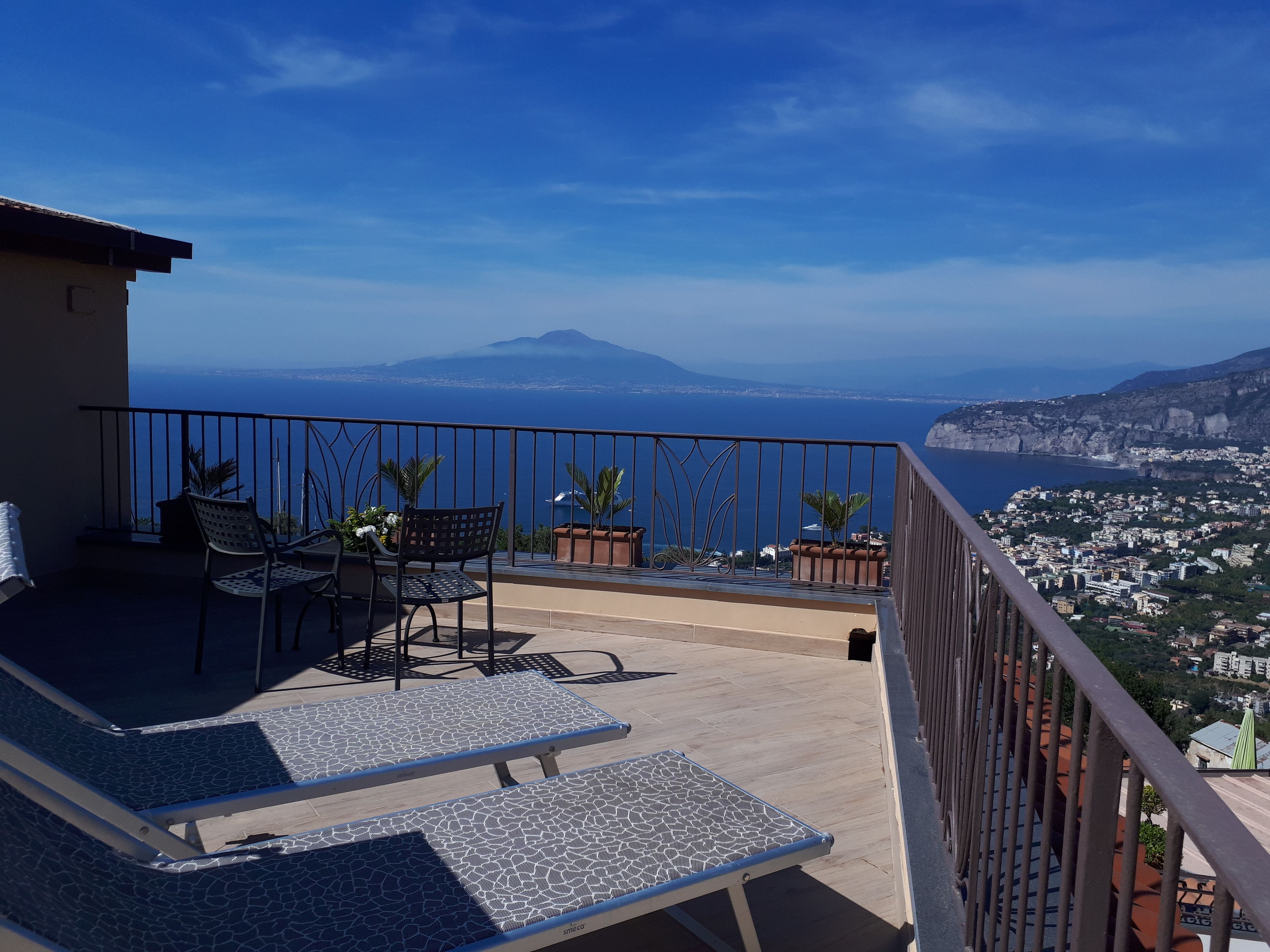 A balcony with two lounge chairs and a table, overlooking a coastal city and a mountain.