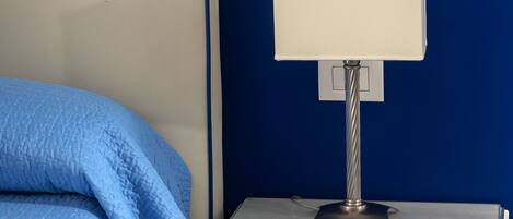 A bed with a blue blanket, a bedside table with a lamp, and a blue wall.