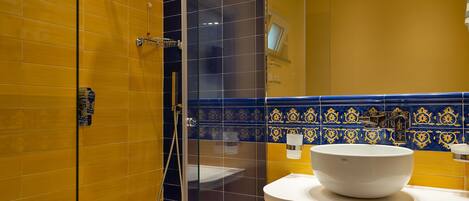 Un baño moderno con baldosas amarillas y azules, una ducha con cerramiento de vidrio, un lavabo blanco con plato y dos toallas colgadas en la pared.