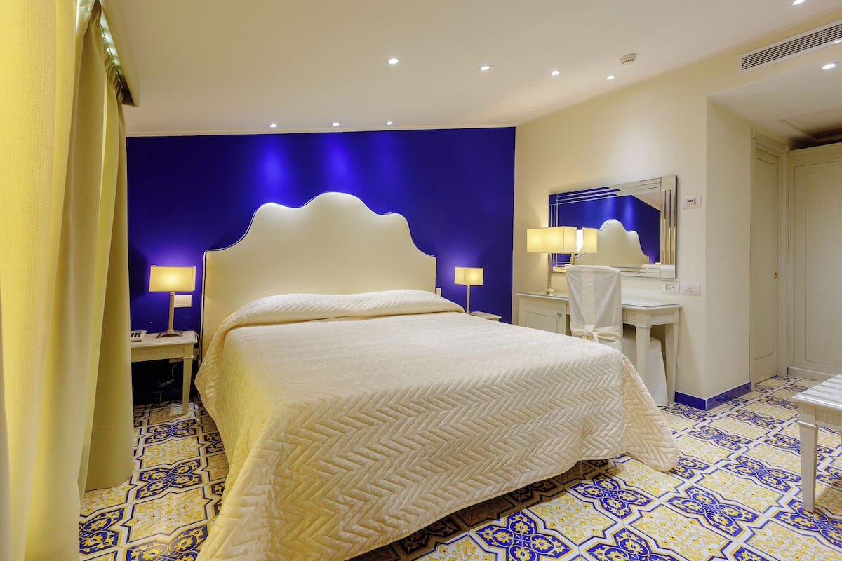 Quarto de hotel com uma cama grande, uma mesinha de cabeceira, um espelho e um espaço de banheiro.