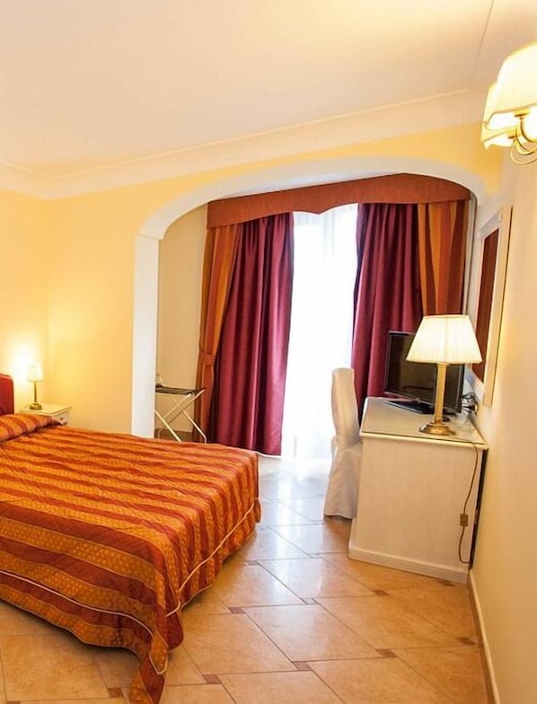 A hotel room with a bed, a desk with a lamp, a chair, and a view through a window with curtains. - La Vue D'Or (Sorrento)