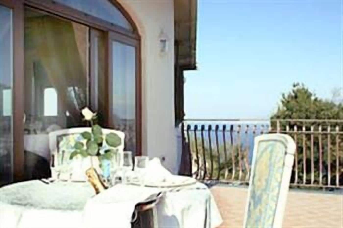 Un balcon avec une table et des chaises, une rose dans un vase et une vue sur la mer.