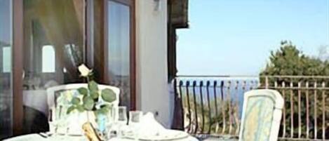 Un balcon avec une table et des chaises, une rose dans un vase et une vue sur la mer.