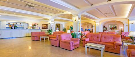 A spacious lobby with pink sofas and armchairs, a reception desk, and a kitchen area in the background.