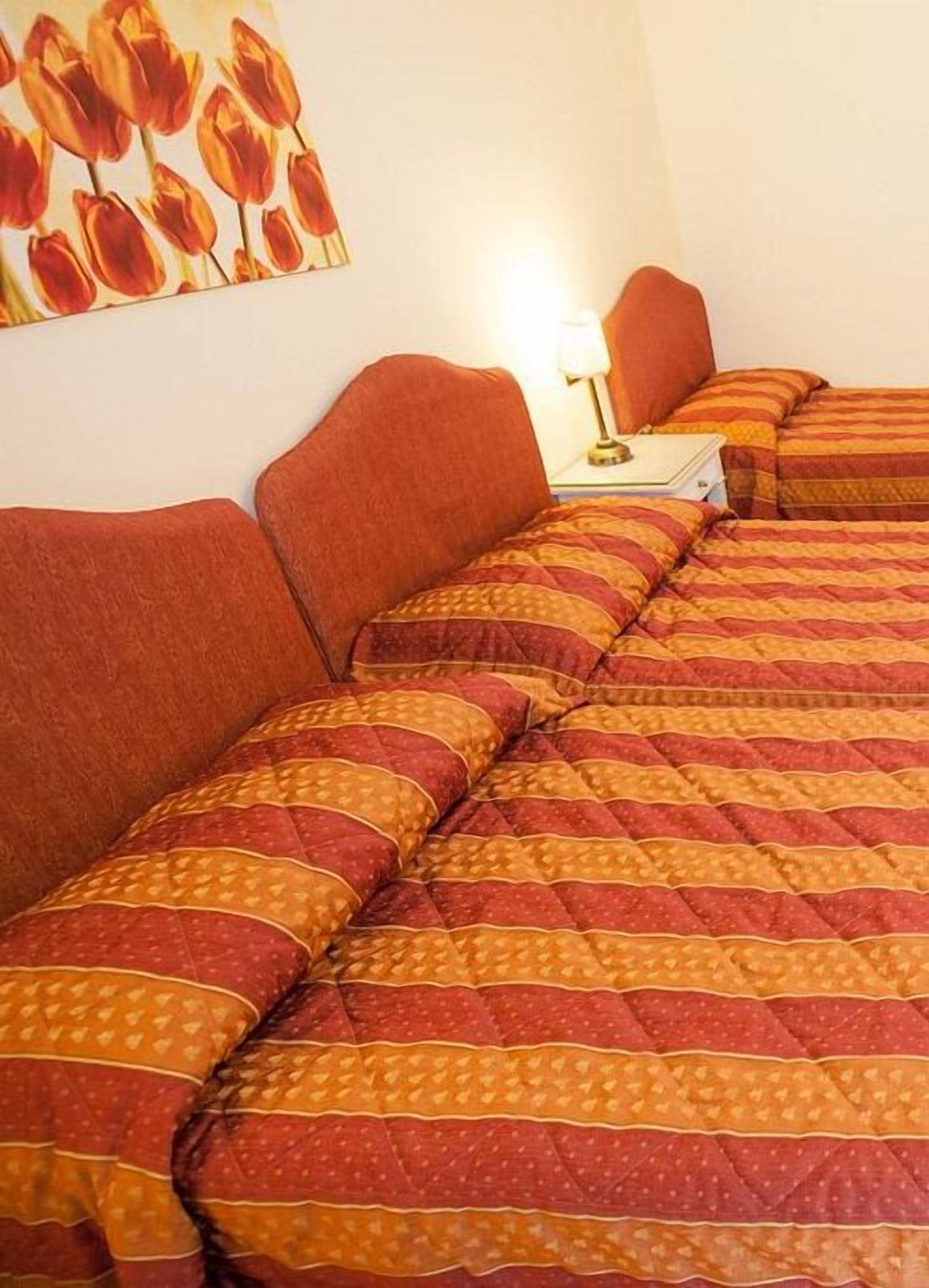 Habitación de hotel con dos camas, cada una con ropa de cama a rayas rojas y anaranjadas. Hay una mesita de noche con una lámpara y una pequeña mesa.
