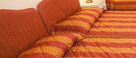 Habitación de hotel con dos camas, cada una con ropa de cama a rayas rojas y anaranjadas. Hay una mesita de noche con una lámpara y una pequeña mesa.