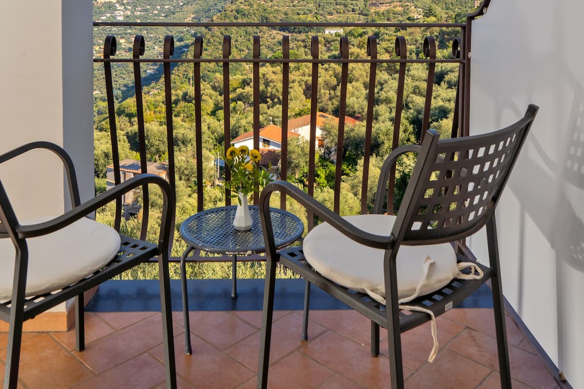 Varanda com duas cadeiras e uma mesa pequena, com vista para uma colina pitoresca com edifícios.