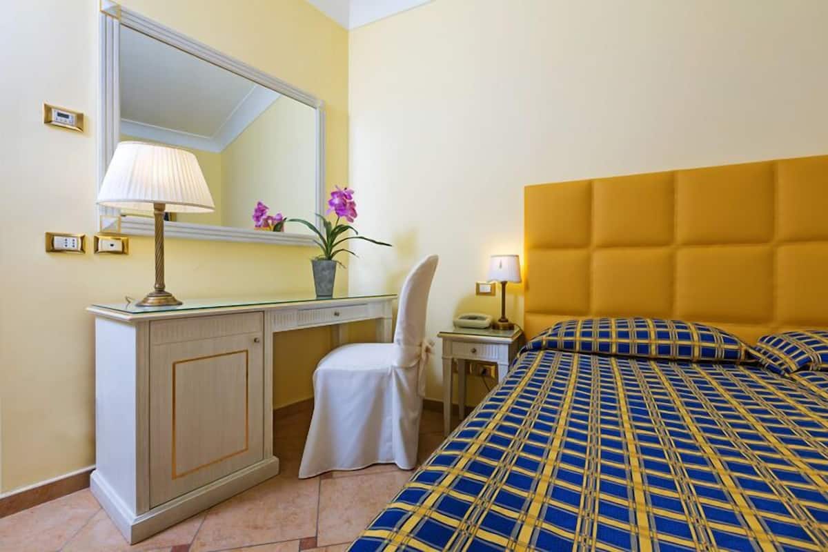 Quarto de hotel com cama, escrivaninha, cadeira, espelho e mesa de cabeceira.