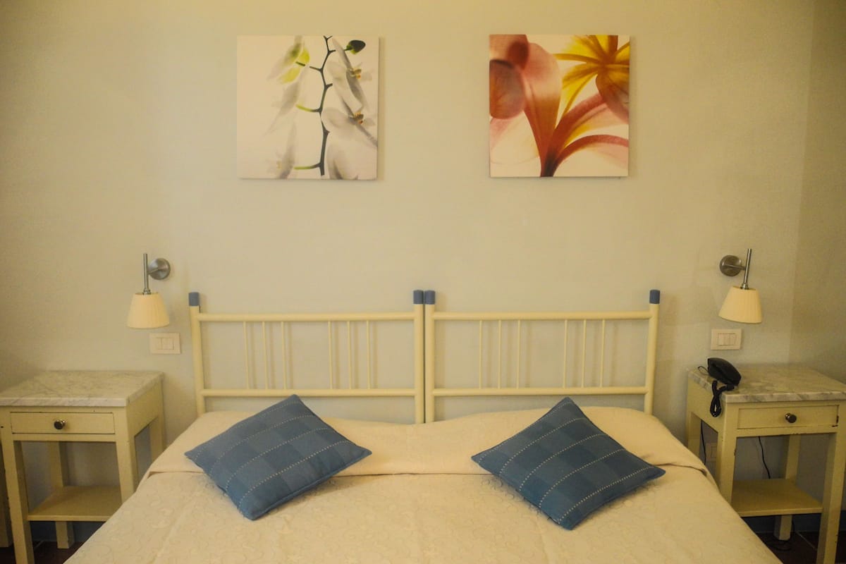 A hotel room with a bed, two pillows, a nightstand with a lamp, and two wall-mounted paintings.