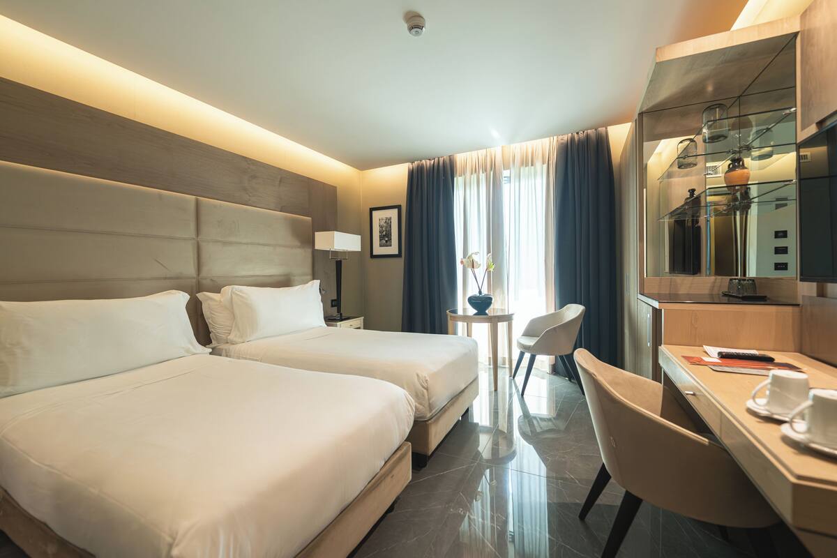 A hotel room with two beds, a desk, a chair, a small table with a vase, and a mirror.