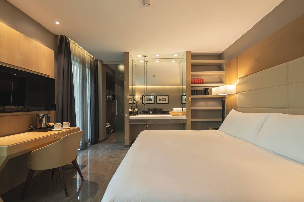A modern hotel room with a large bed, a desk, a chair, a TV, a bathroom, and a bookshelf.