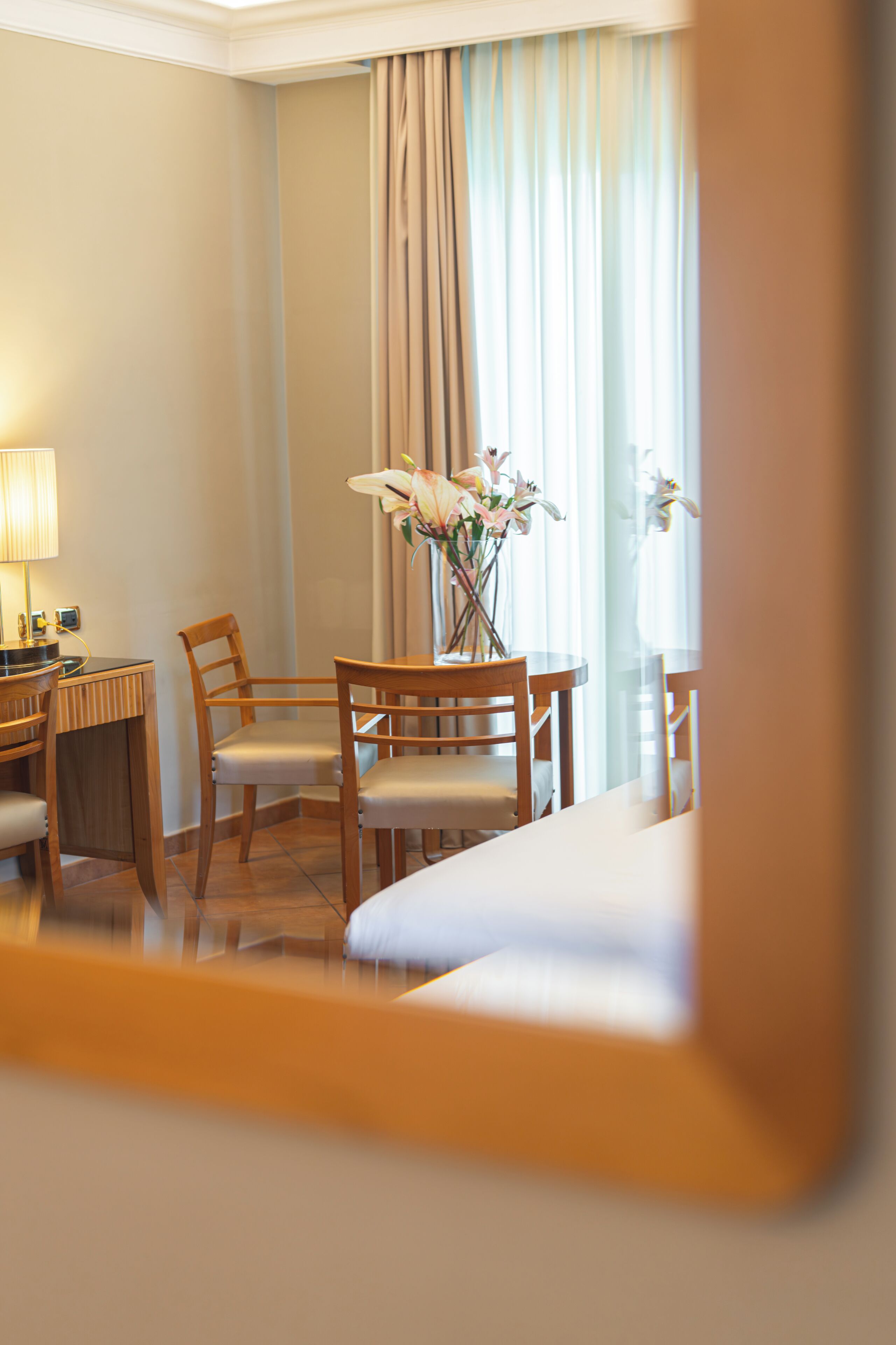 Une chambre d’hôtel avec un lit, un bureau doté d’une lampe, deux chaises et un vase avec des fleurs.