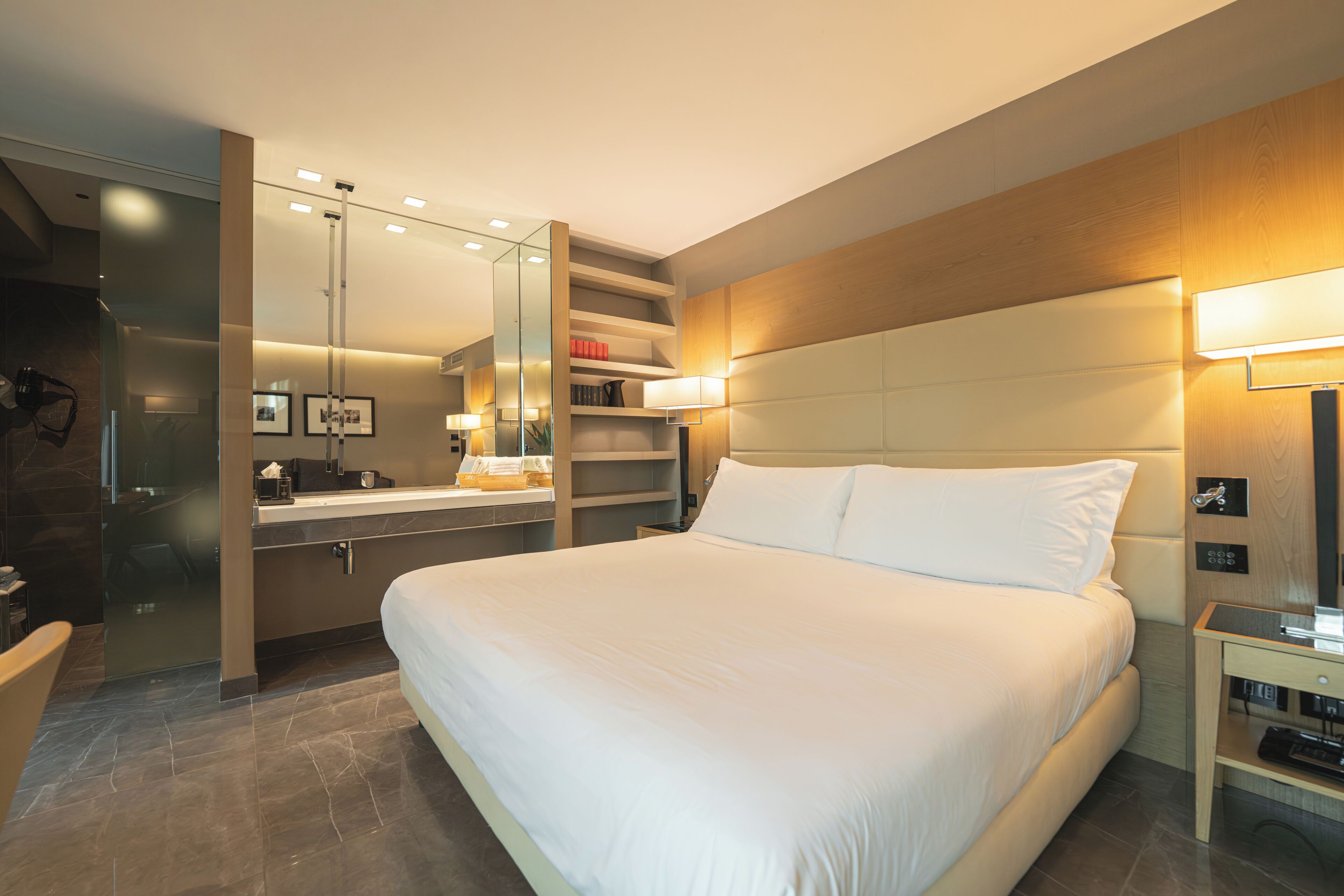 A modern hotel room with a large bed, a vanity with a mirror, a bedside table, and a built-in shelf with books.