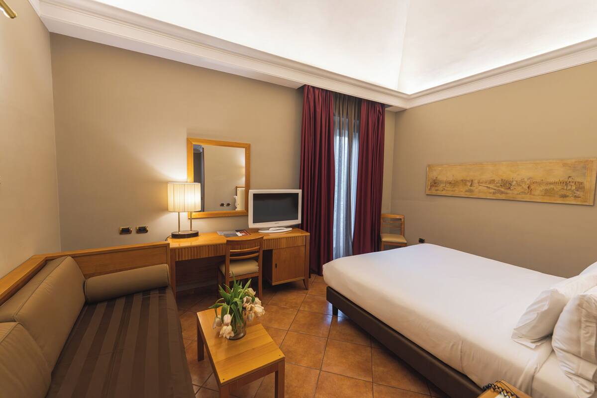 A hotel room with a bed, desk, chair, TV, and a painting on the wall.