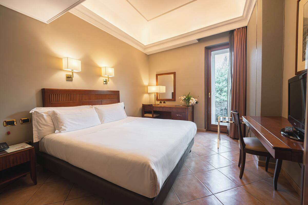 A hotel room with a large bed, bedside lamps, a desk with a chair, a TV, and a window with curtains.