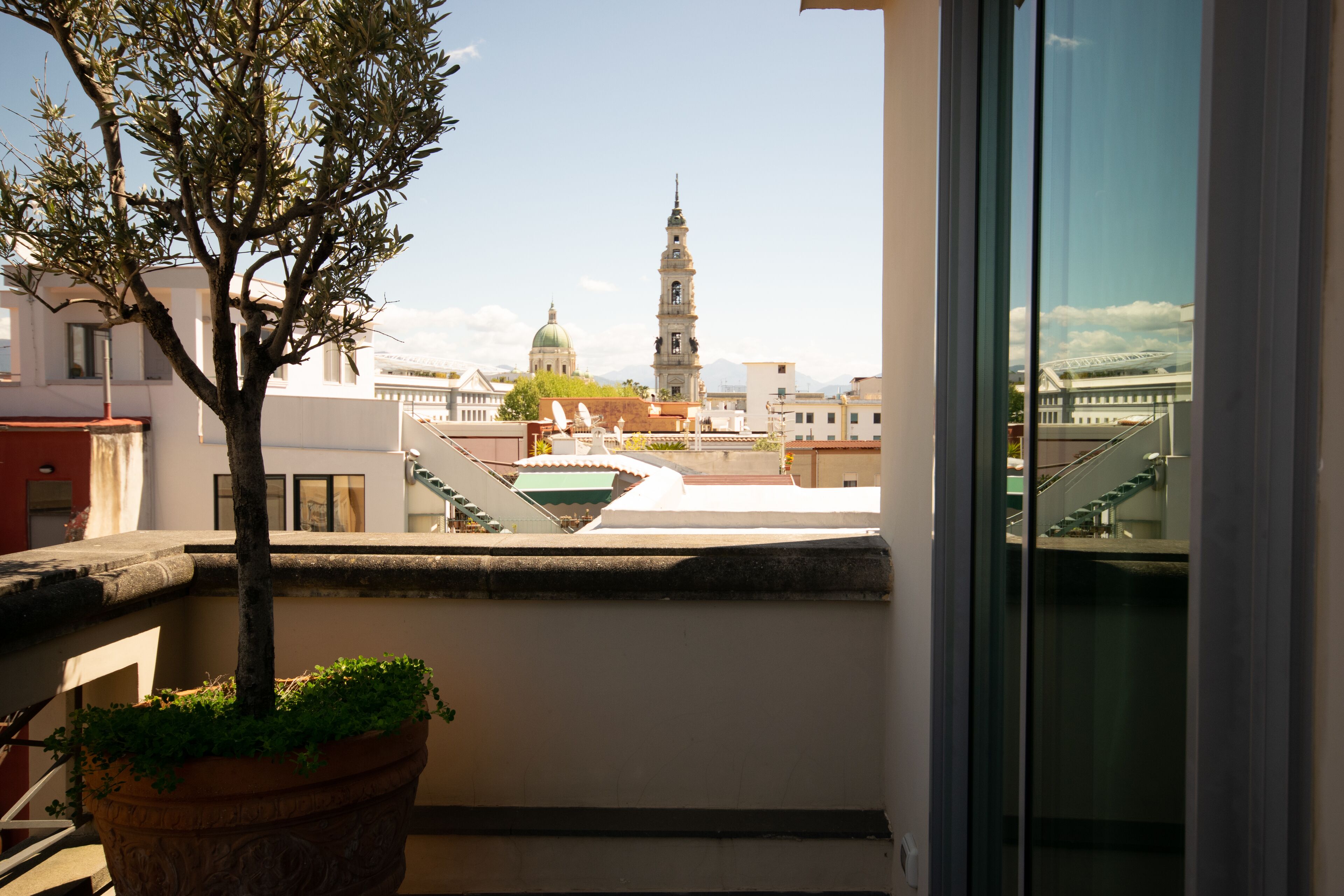 Ein Balkon mit einem Blumentopf und einem Baum, mit Blick auf eine Stadtlandschaft, die durch einen markanten Turm und ein Kuppelgebäude geprägt ist.