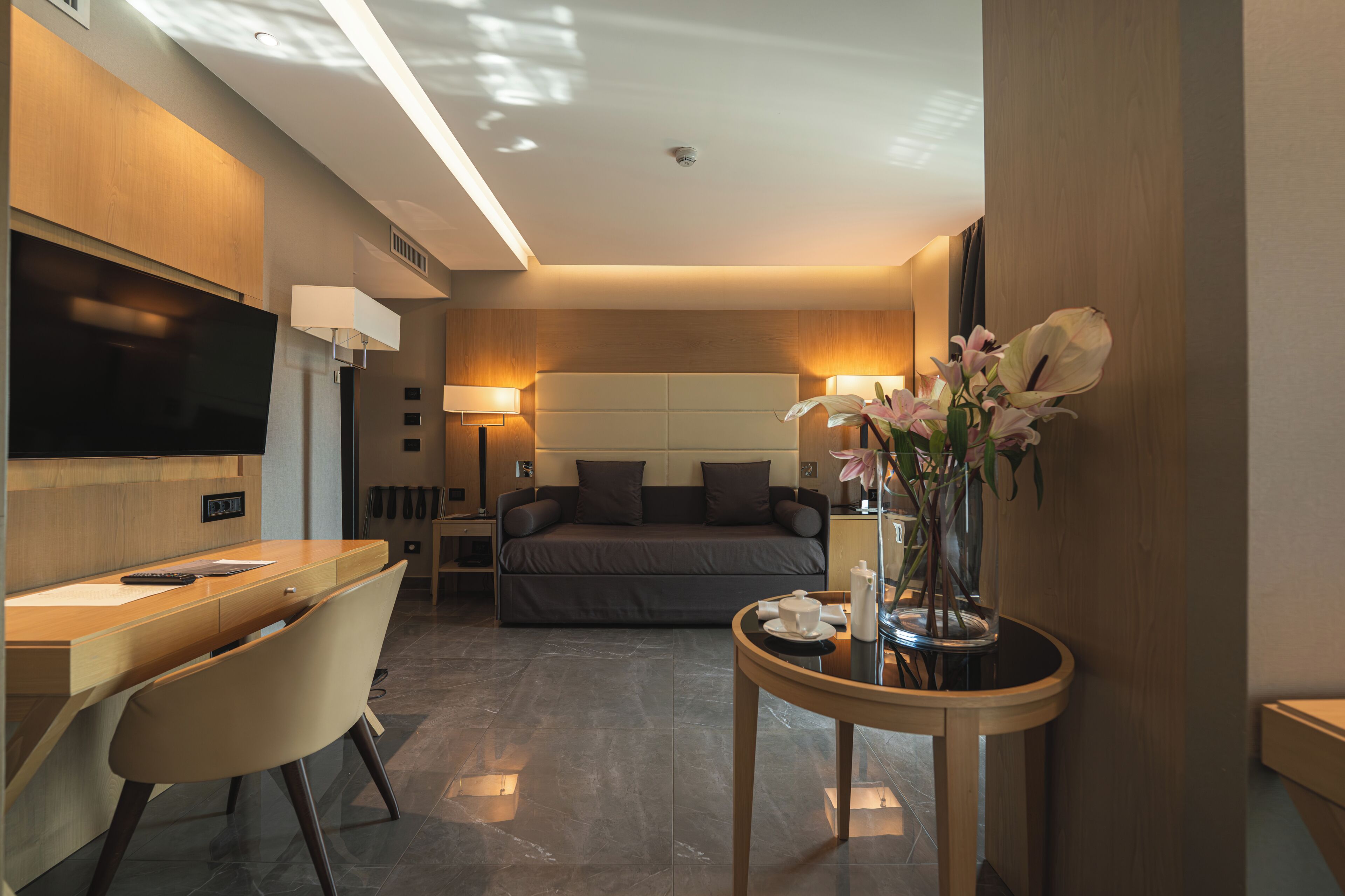 Una habitación de hotel moderna con cama, escritorio, silla, televisor, lámparas de noche y un jarrón con flores.