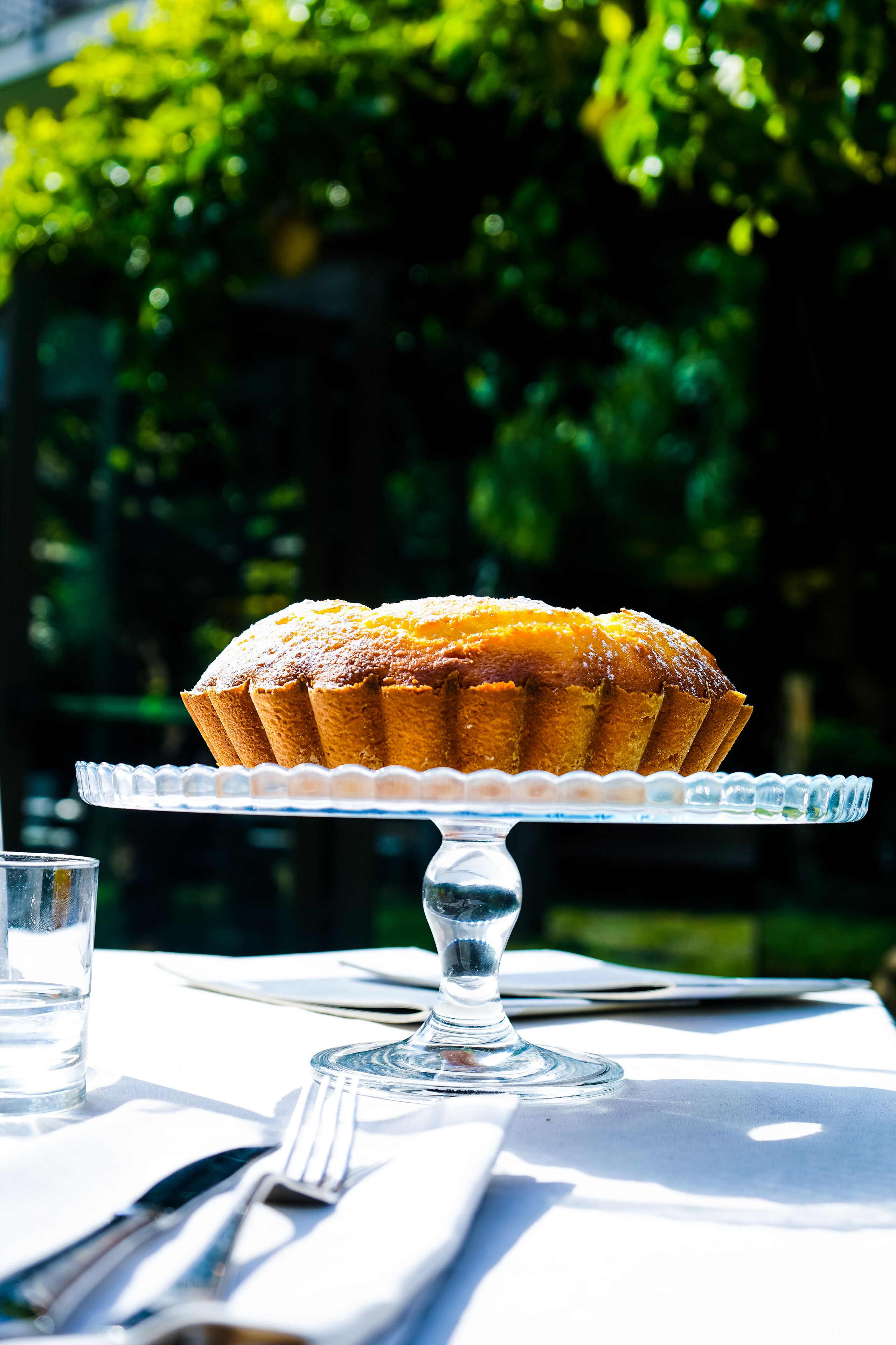 A cake on a glass stand on a table with a white tablecloth.