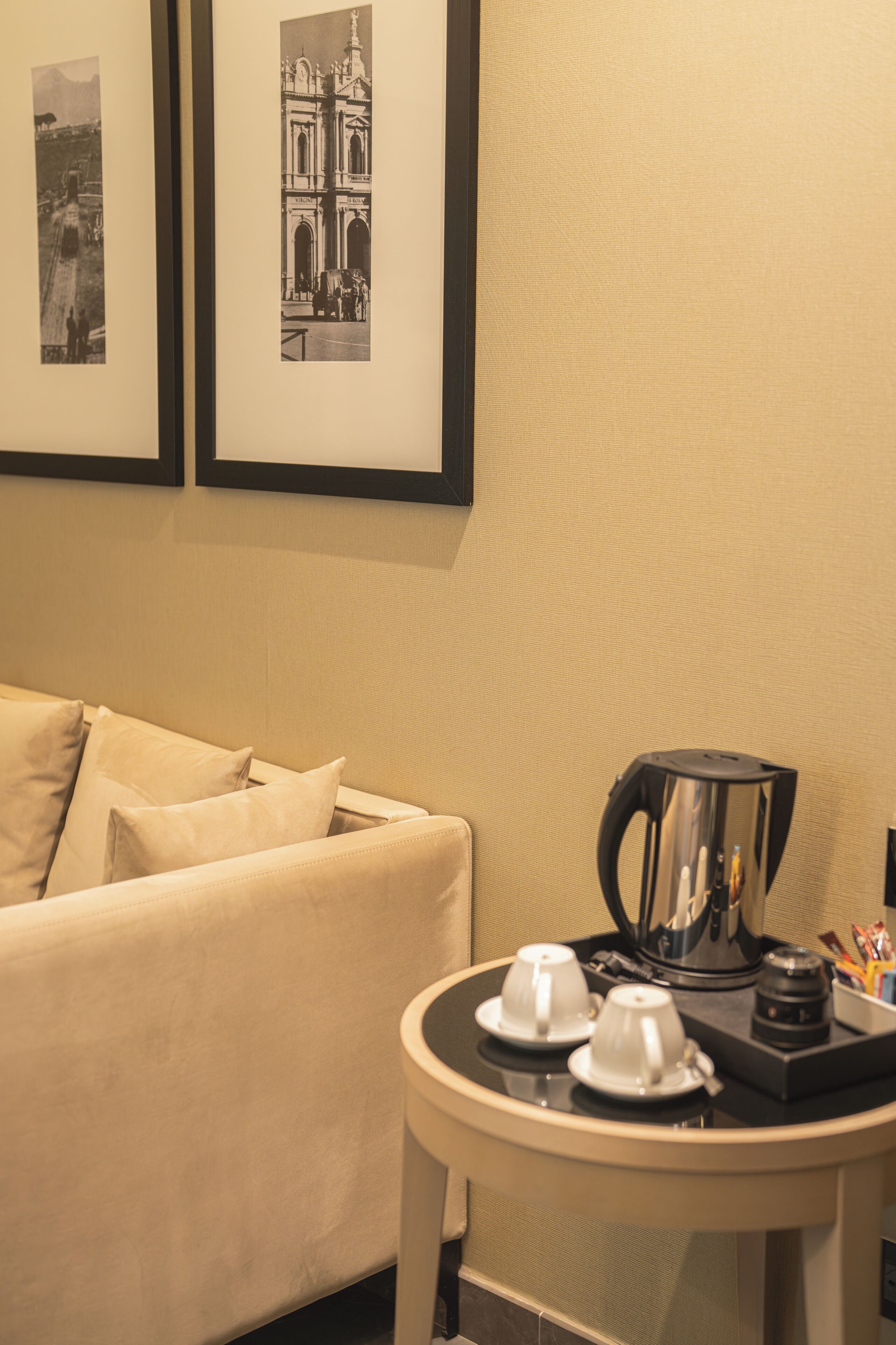 Quarto de hotel com sofá, uma mesinha redonda com cafeteira, chávenas e quadros nas paredes.