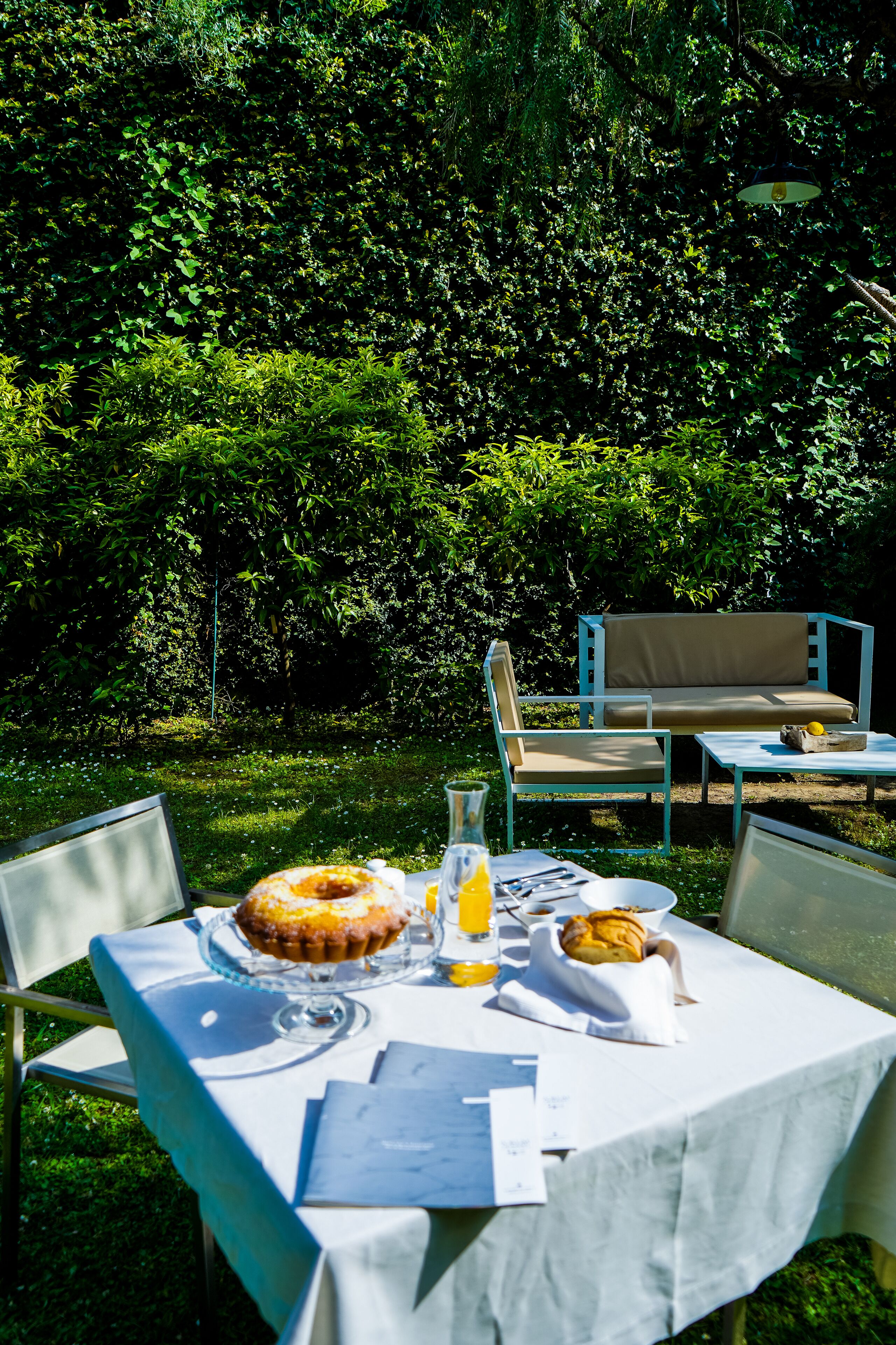 An outdoor dining area with a table set for breakfast, including a cake and a basket of bread, surrounded by lush greenery.