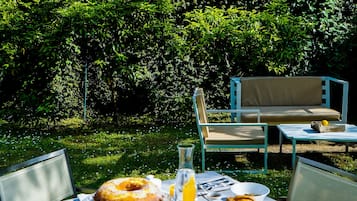 An outdoor dining area with a table set for breakfast, including a cake and a basket of bread, surrounded by lush greenery.