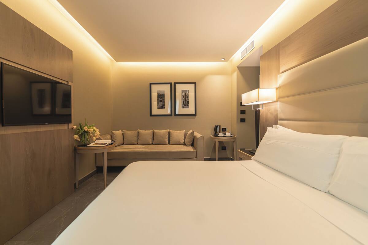 A hotel room with a bed, a sofa, a small table, a lamp, and two framed pictures on the wall.