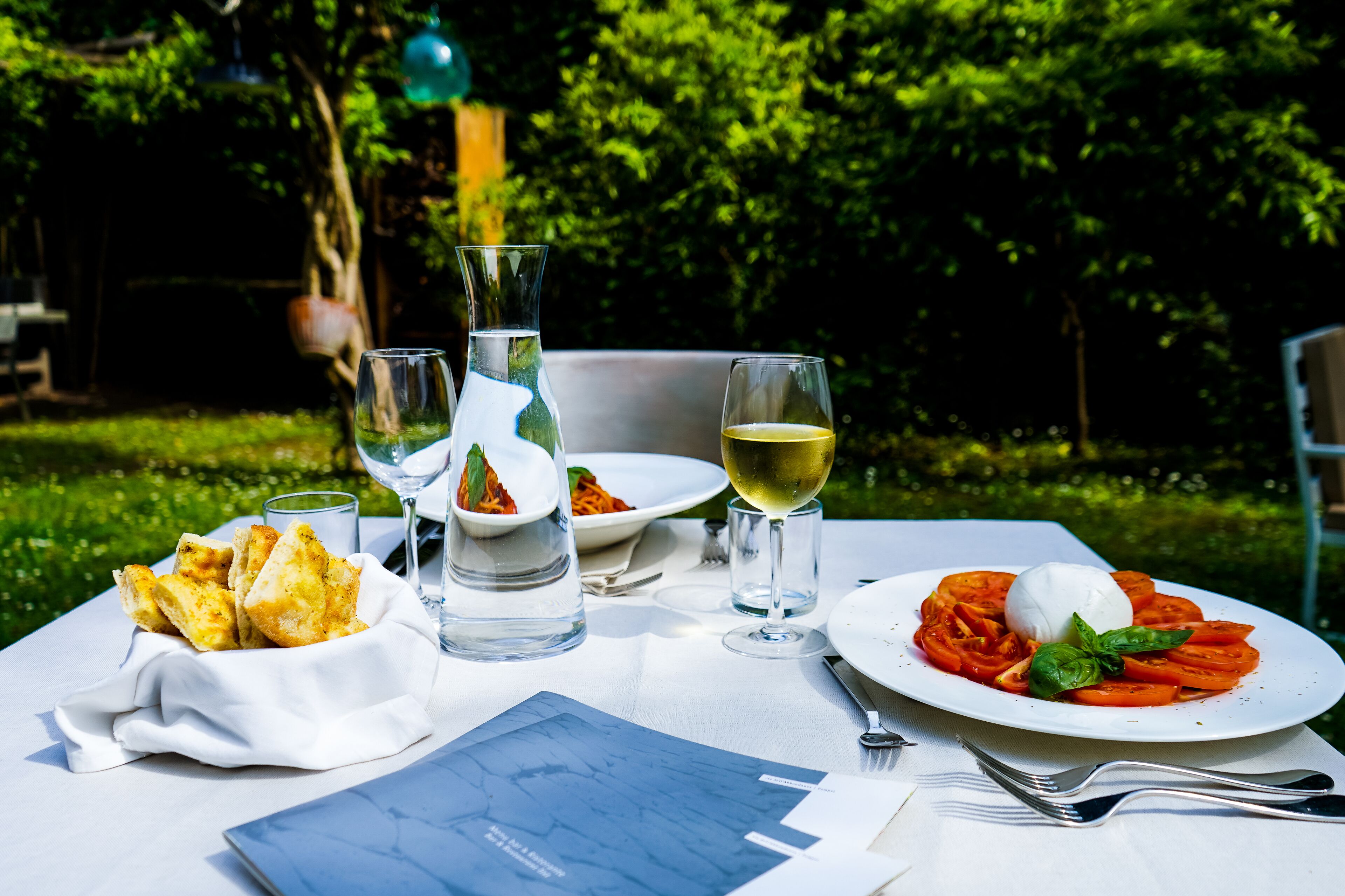 Una mesa al aire libre con un plato de comida, una copa de vino blanco y una botella de agua.
