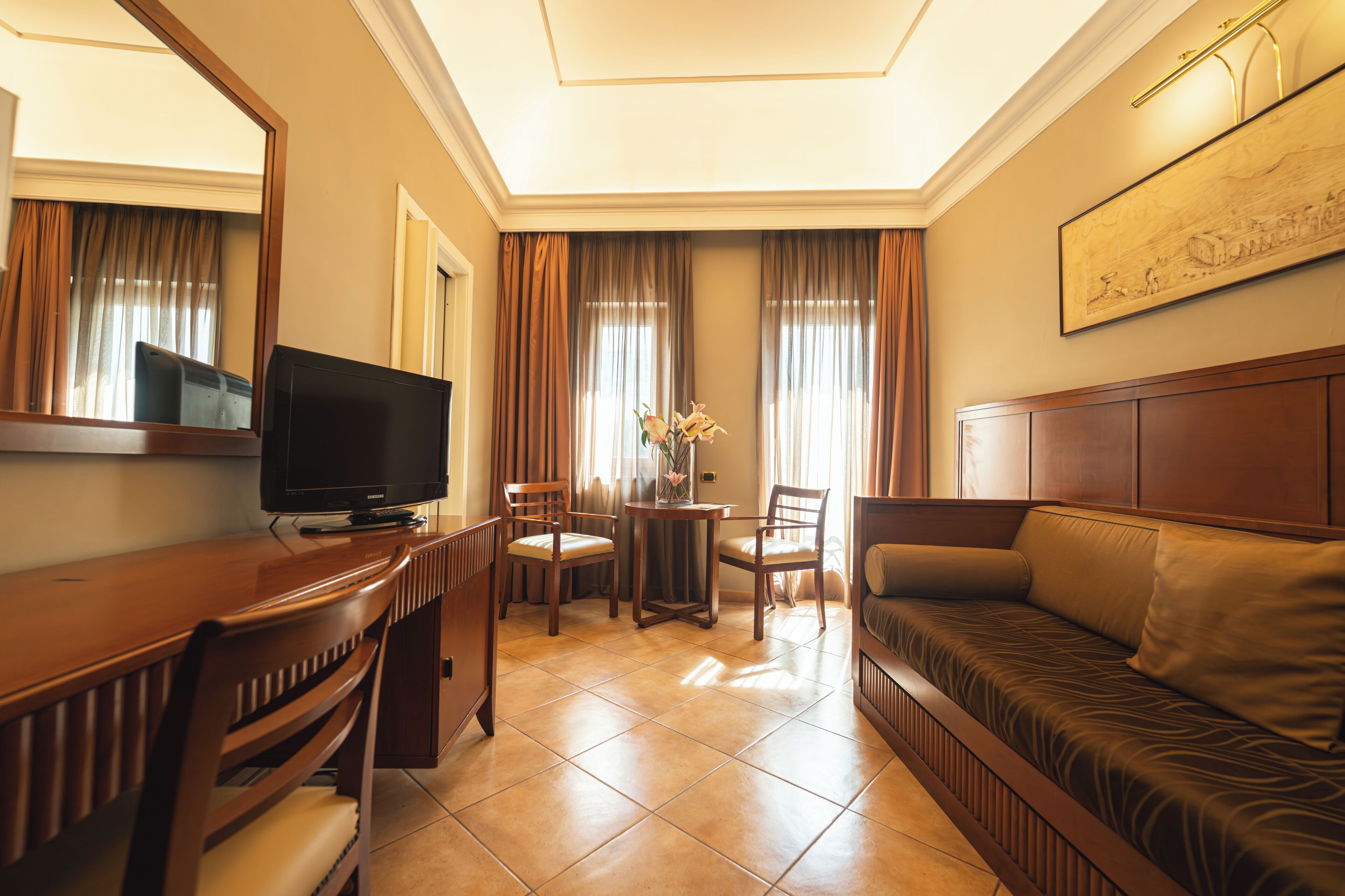 Habitación de hotel con televisión, sofá, mesa de comedor con sillas y un florero con flores.