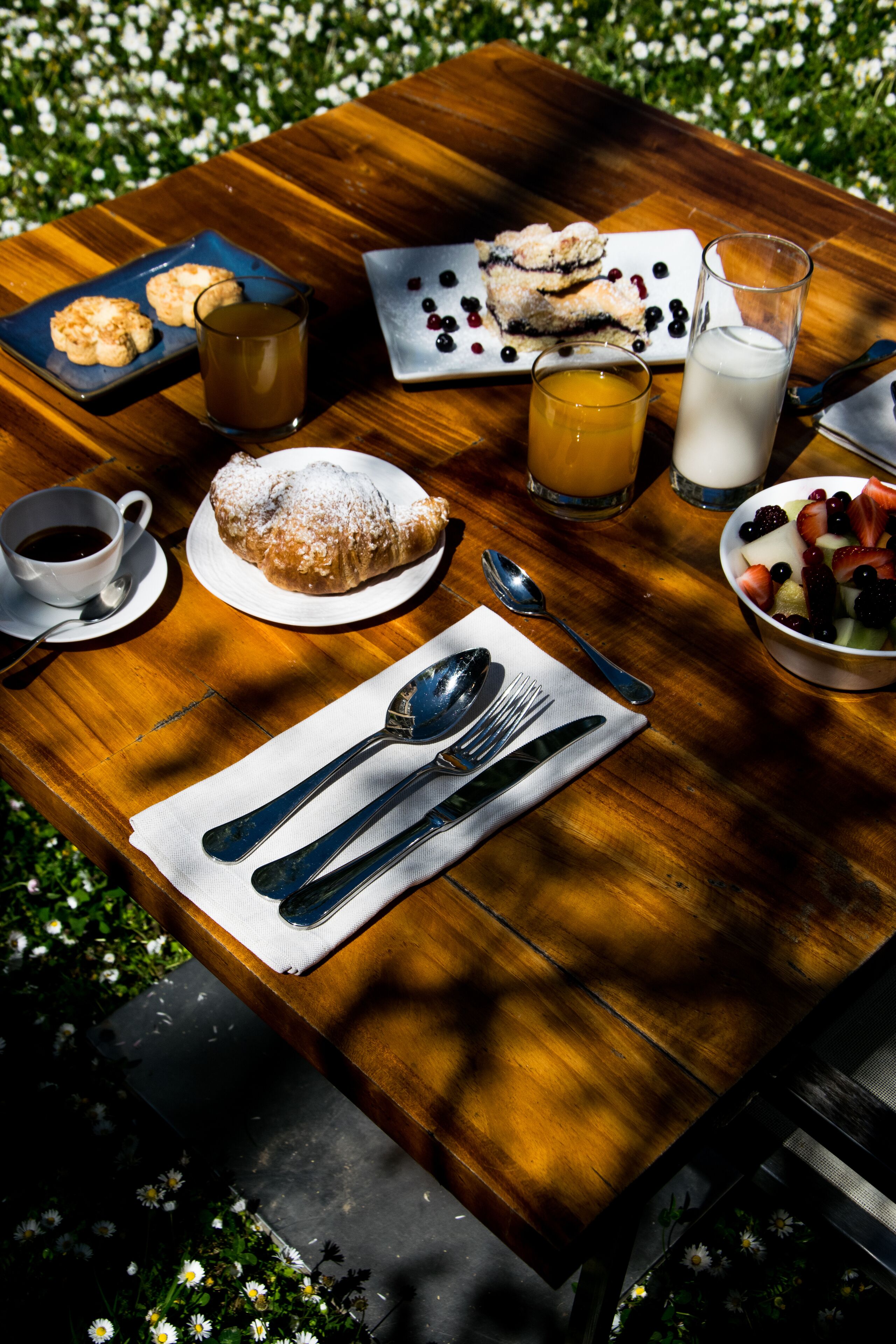 A wooden table with a meal setup including a plate of pastries, a glass of orange juice, a cup of coffee, and a bowl of fruit.