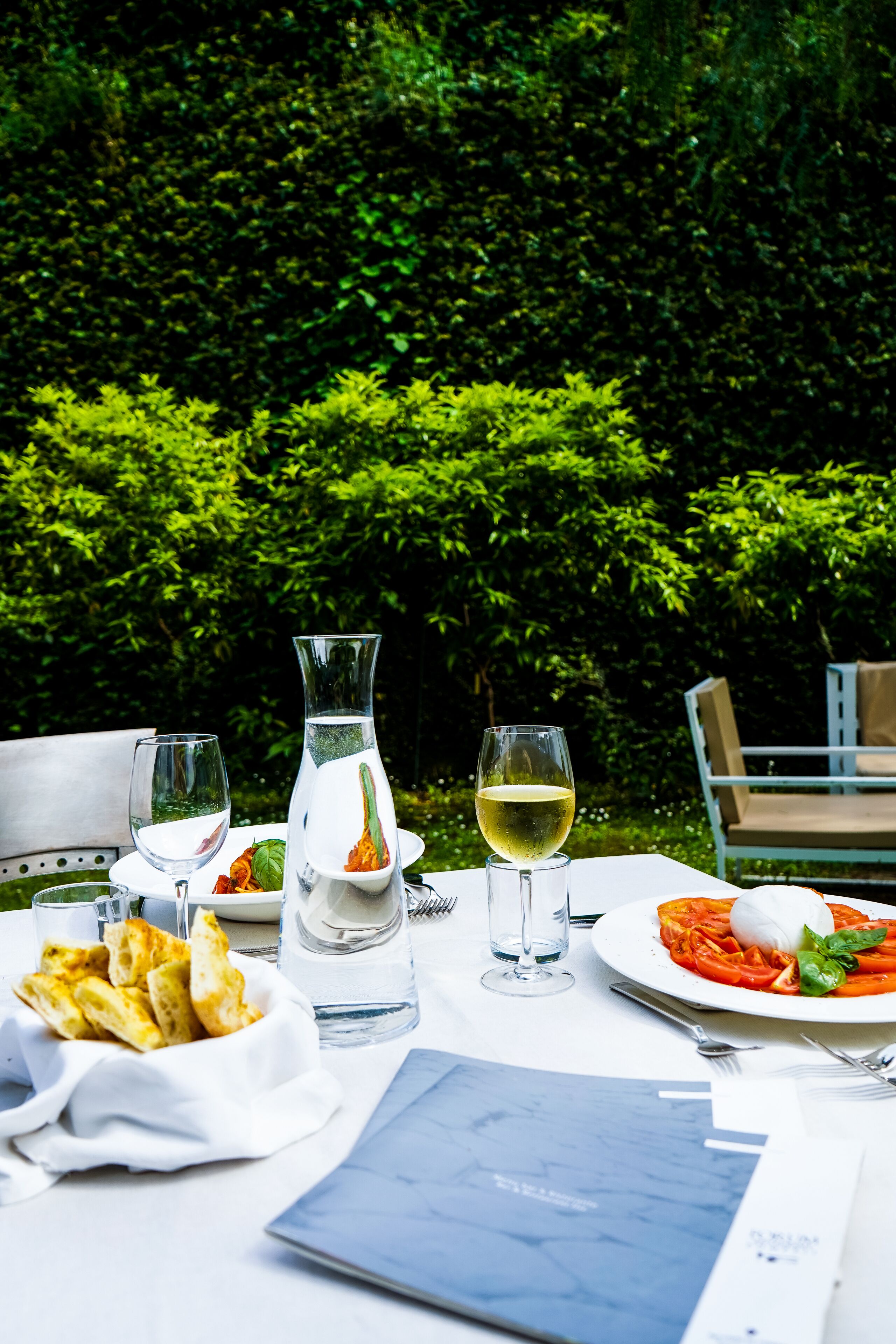Una mesa de comedor al aire libre con una jarrón, copas de vino, un plato de pasta y una cesta de pan.