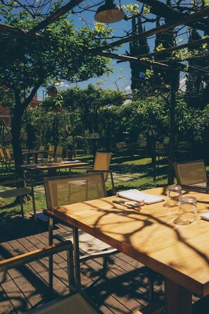 Un área de comedor al aire libre con mesas y sillas de madera, rodeada de árboles y follaje.
