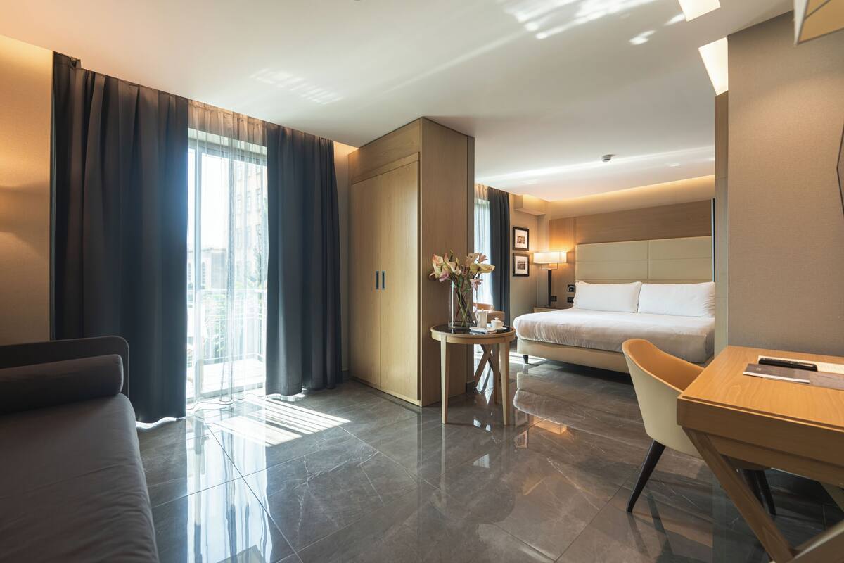 A hotel room with a bed, a desk, a chair, a small table with a vase, and a wardrobe.