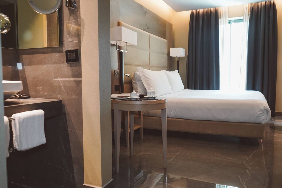 A hotel room with a bed, a bedside table, a mirror, a lamp, and a window with curtains.