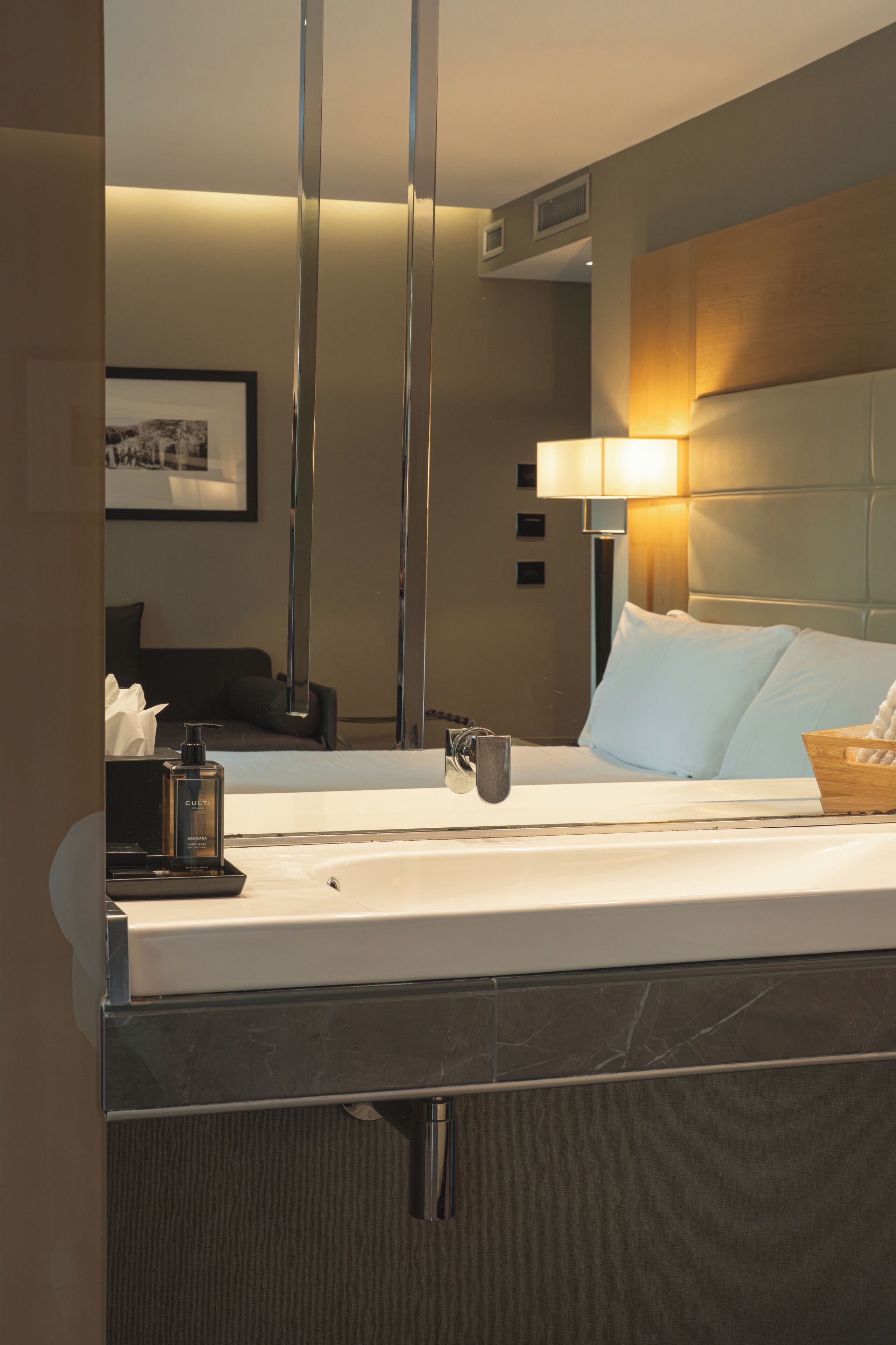 A hotel room with a large mirror, a bed with white linens, a bedside lamp, a small table with a tissue box, and a framed picture on the wall.