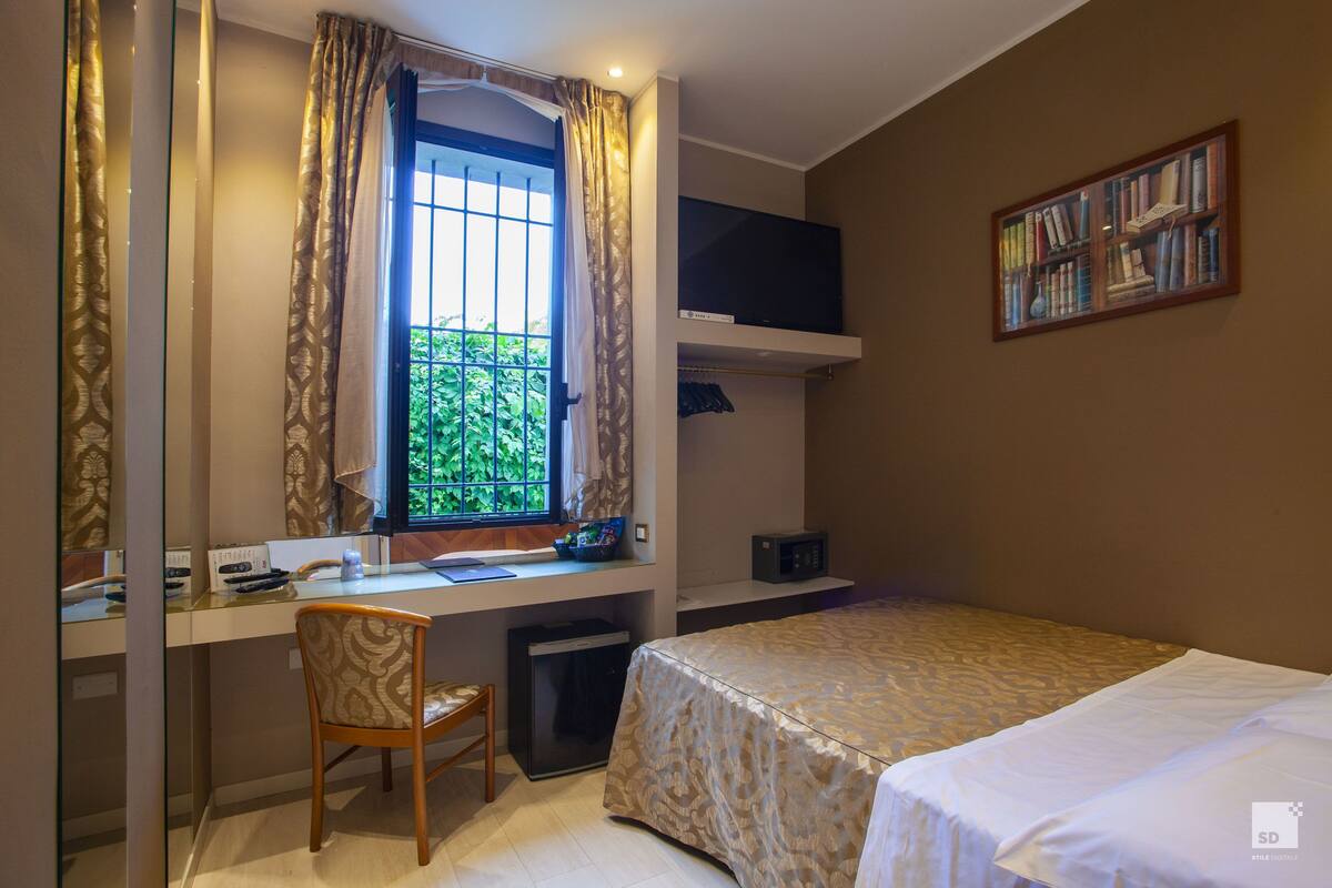 Habitación de hotel pequeña con cama, escritorio con silla, televisor y estantería.