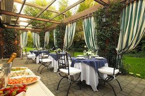 Een buiten eethoek met tafels gedekt voor een maaltijd, overdekt door een pergola met groen-wit gestreepte gordijnen.