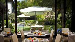 An outdoor dining area with tables set for breakfast, surrounded by greenery and large umbrellas. - Savoia Hotel Country House (Bologna)