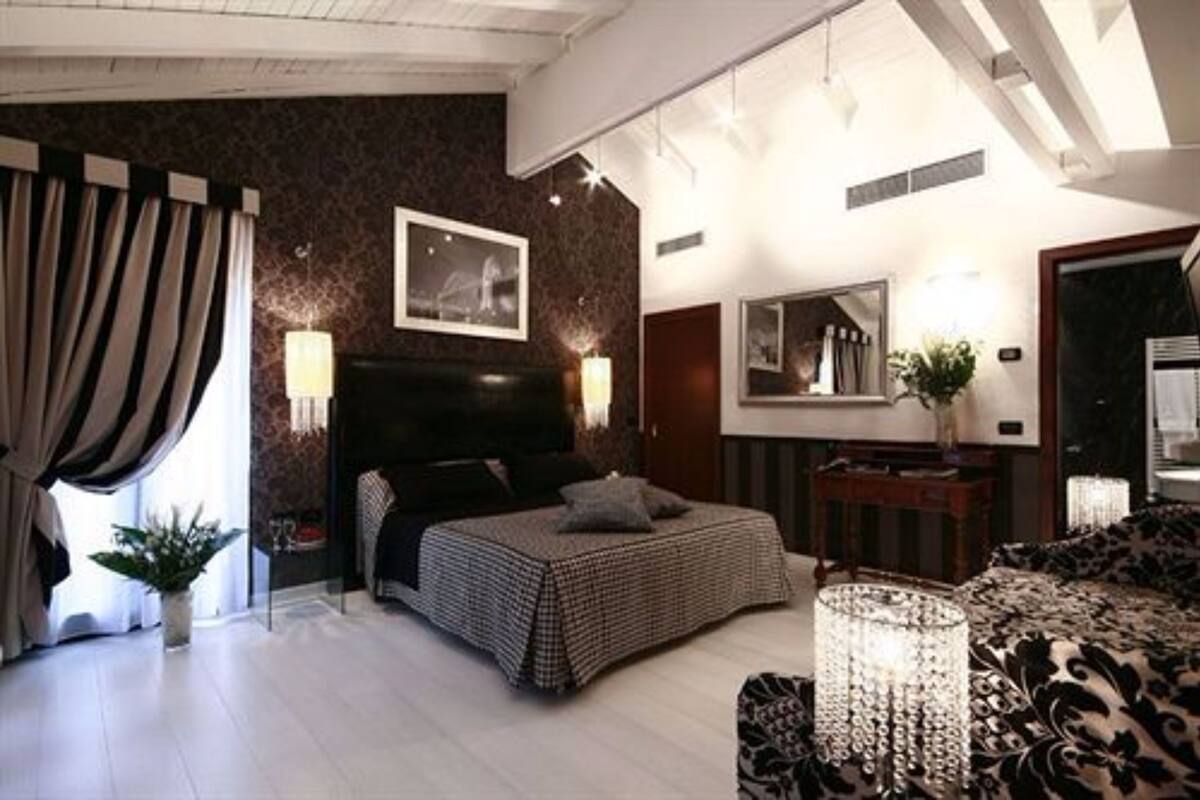 Un dormitorio con una cama grande, un cabecero oscuro, una mesita de noche con lámpara, un espejo y una ventana con cortinas.