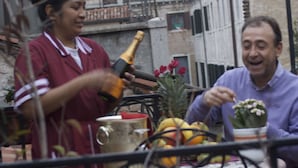 Iemand houdt een fles vast, een tafel met fruit en een bloempot.