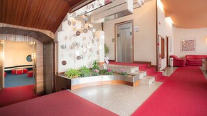 A modern lobby with a red carpet, a central planter with flowers, a staircase, and a red sofa. - Albergo Residence Italia (Pordenone)