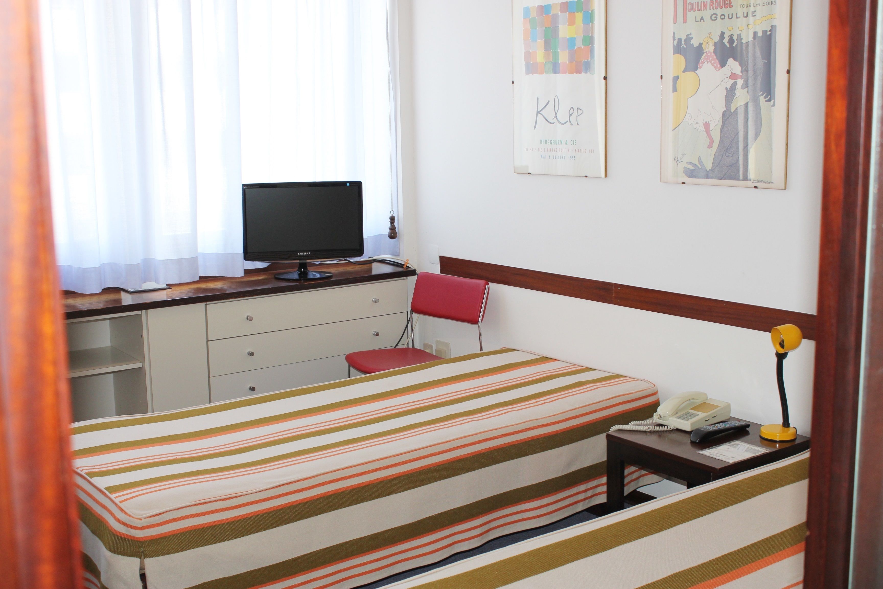 Un dormitorio con cama, escritorio con computadora, silla, teléfono y dos carteles en la pared.
