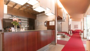 A hotel reception area with a wooden reception desk, a red carpet, and a seating area with red sofas. - Albergo Residence Italia (Pordenone)