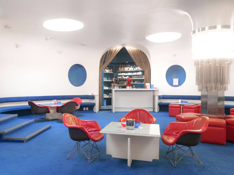 A modern lounge area with blue carpet, red and black chairs, a white table, and a bar area with shelves of bottles.