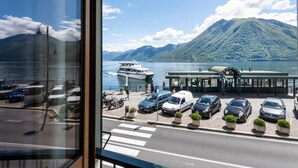 Une vue depuis une fenêtre donnant sur le bord de l’eau, avec un quai pour bateaux, des voitures garées devant et une montagne en arrière-plan.