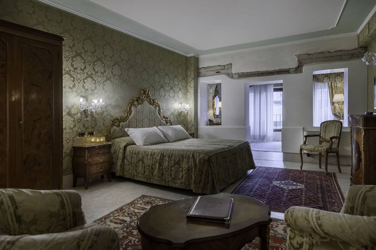 A bedroom with a large bed, a nightstand, a dresser, and a chair. There is a window with curtains and a view of the outside.
