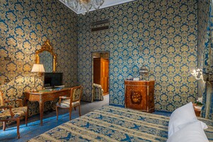 Una stanza con carta da parati decorata, una scrivania con una lampada, una sedia, un letto e un armadietto in legno.