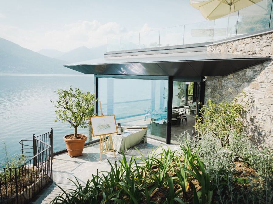 A lakeside terrace with a table and chairs, a potted plant, and a painting on easels.