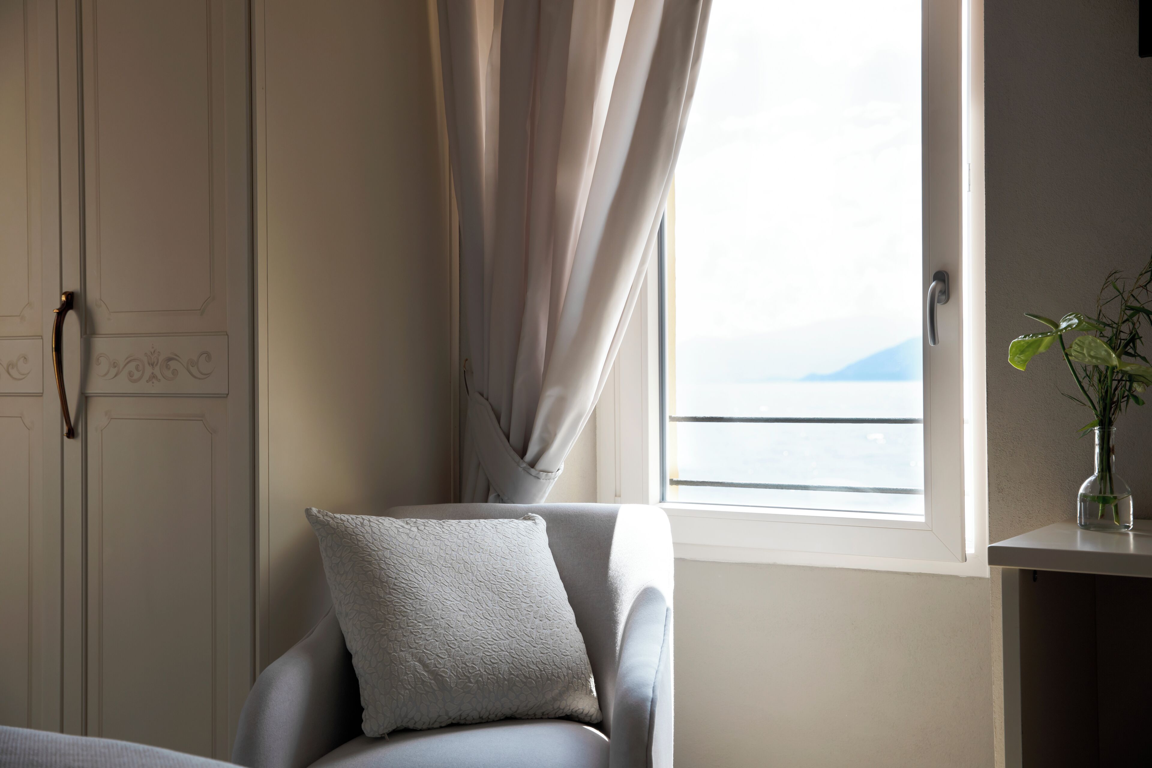 Una habitación con vistas al mar a través de una ventana, una silla con cojín y un florero con una planta sobre una mesa.