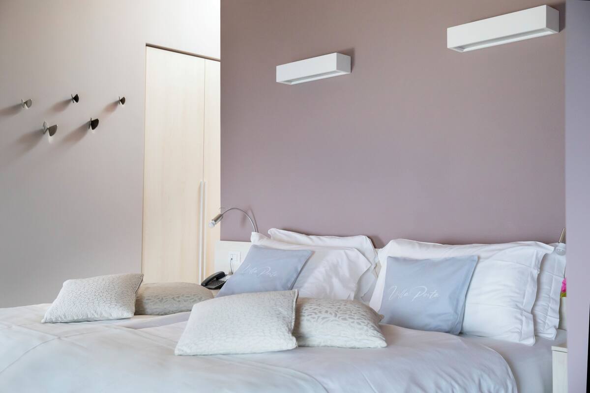 Ein ordentlich bezogenes Bett mit weißen und hellblauen Kissen, ein Nachttisch mit Lampe sowie eine Wandleuchte.
