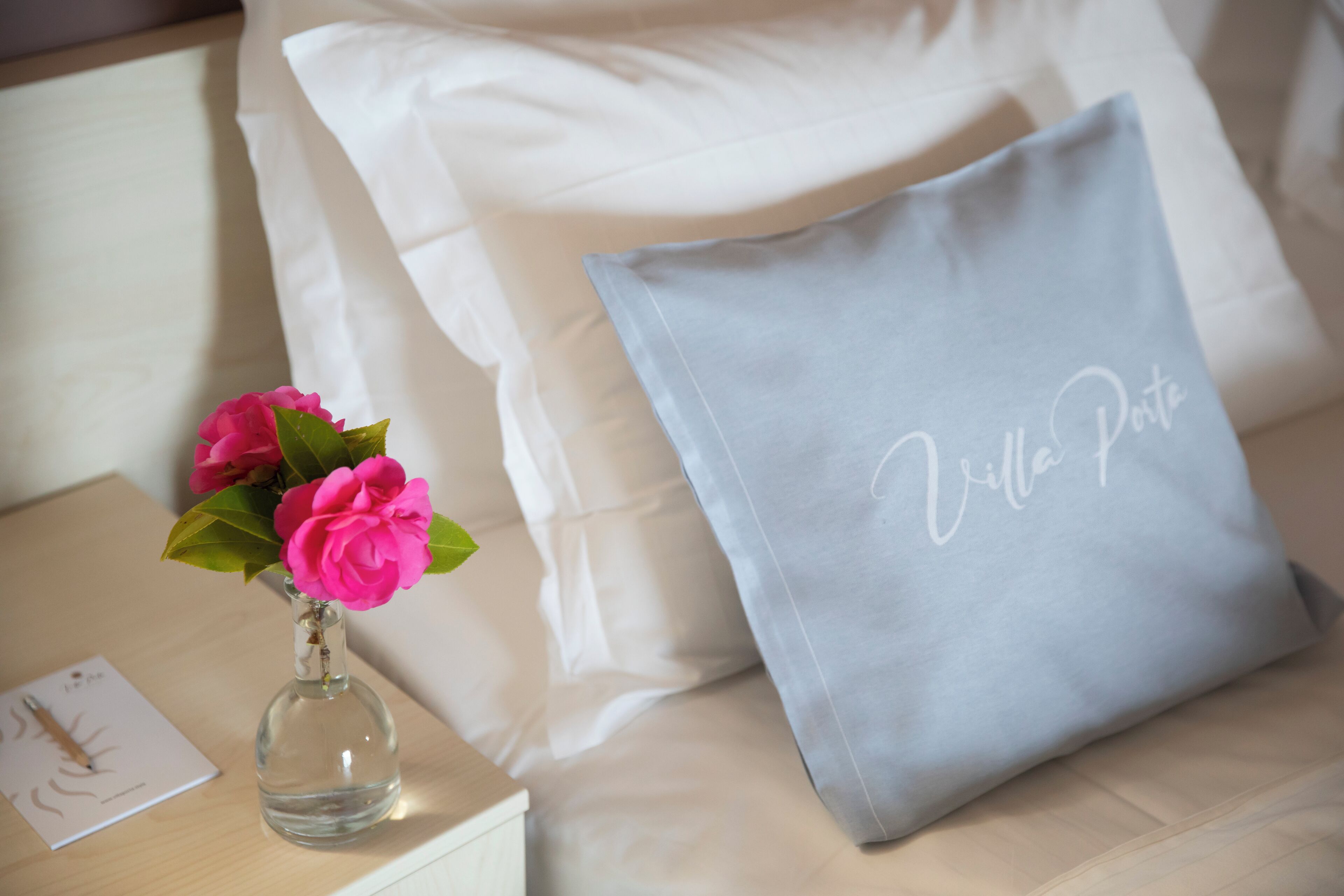 A bed with white pillows and a grey pillow with "Villa Porto" embroidered on it. A clear glass vase with pink flowers is on a bedside table.